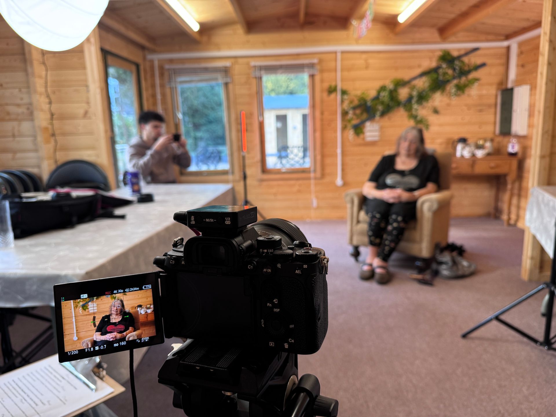 A camera on a tripod records a person sitting in a chair inside a wood-paneled room.