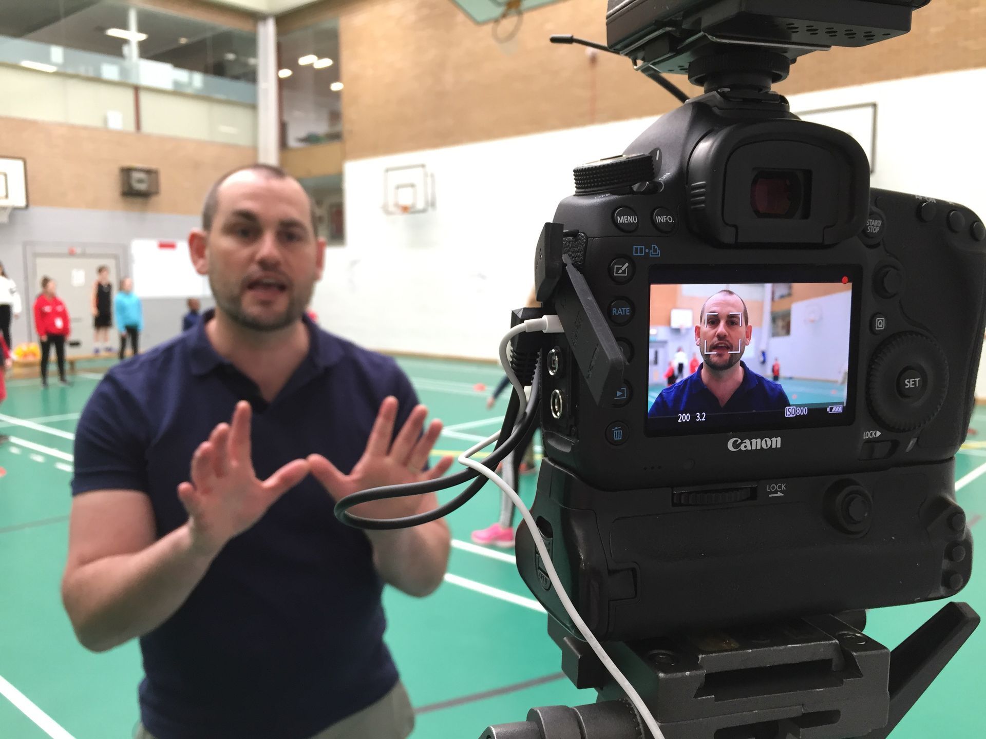A person speaks to a camera on a tripod in a sports hall, visible on the camera’s digital viewfinder.