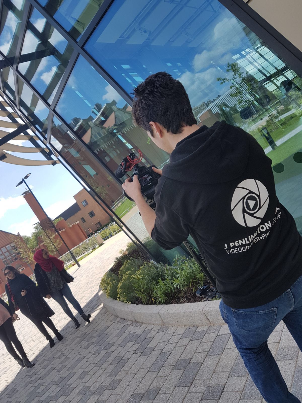 A videographer filming two people outdoors in front of a modern building with glass walls.
