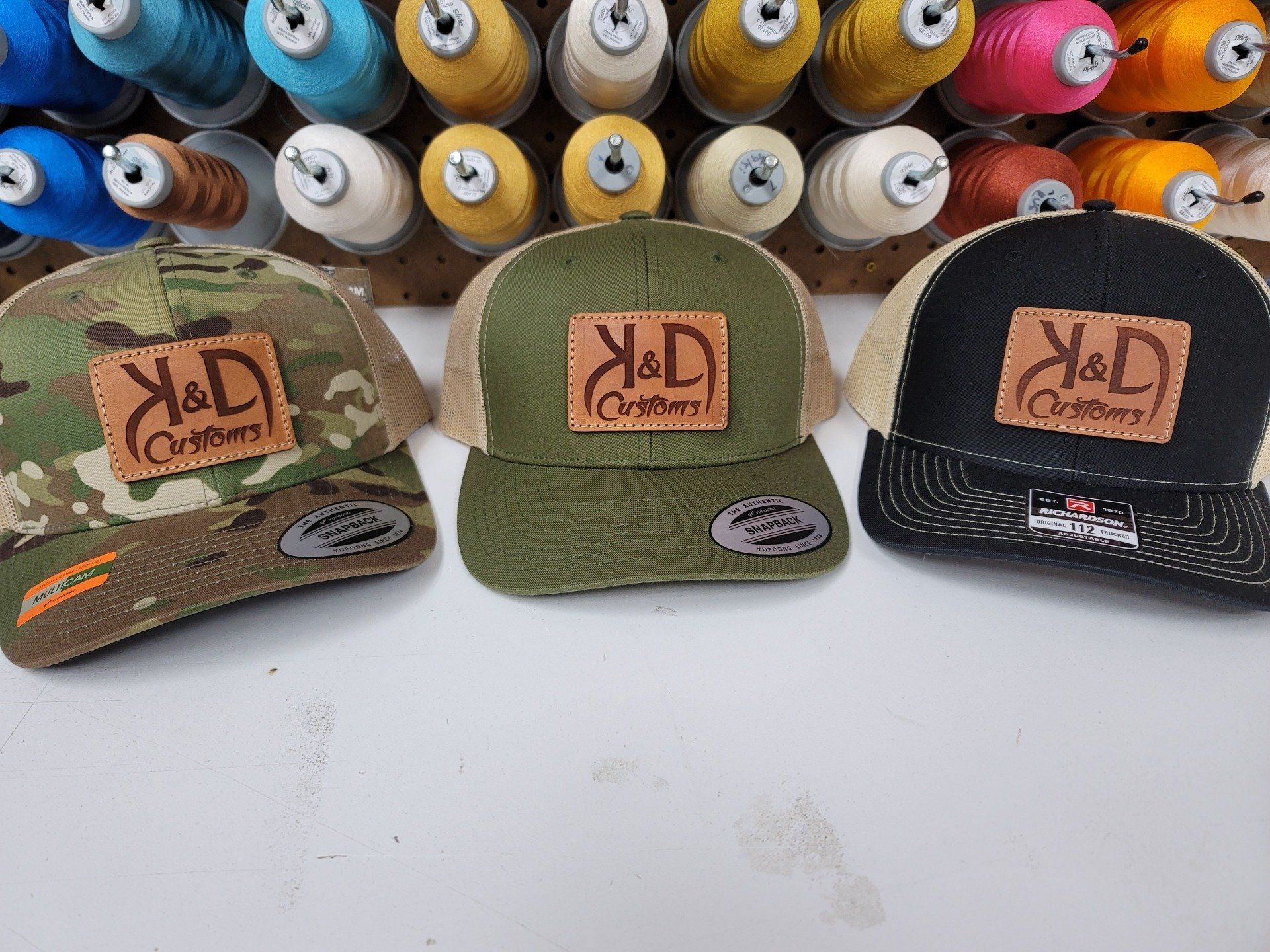 Three trucker hats with leather patches, K&D Outdoors logo, in camouflage, olive, and black with tan mesh.