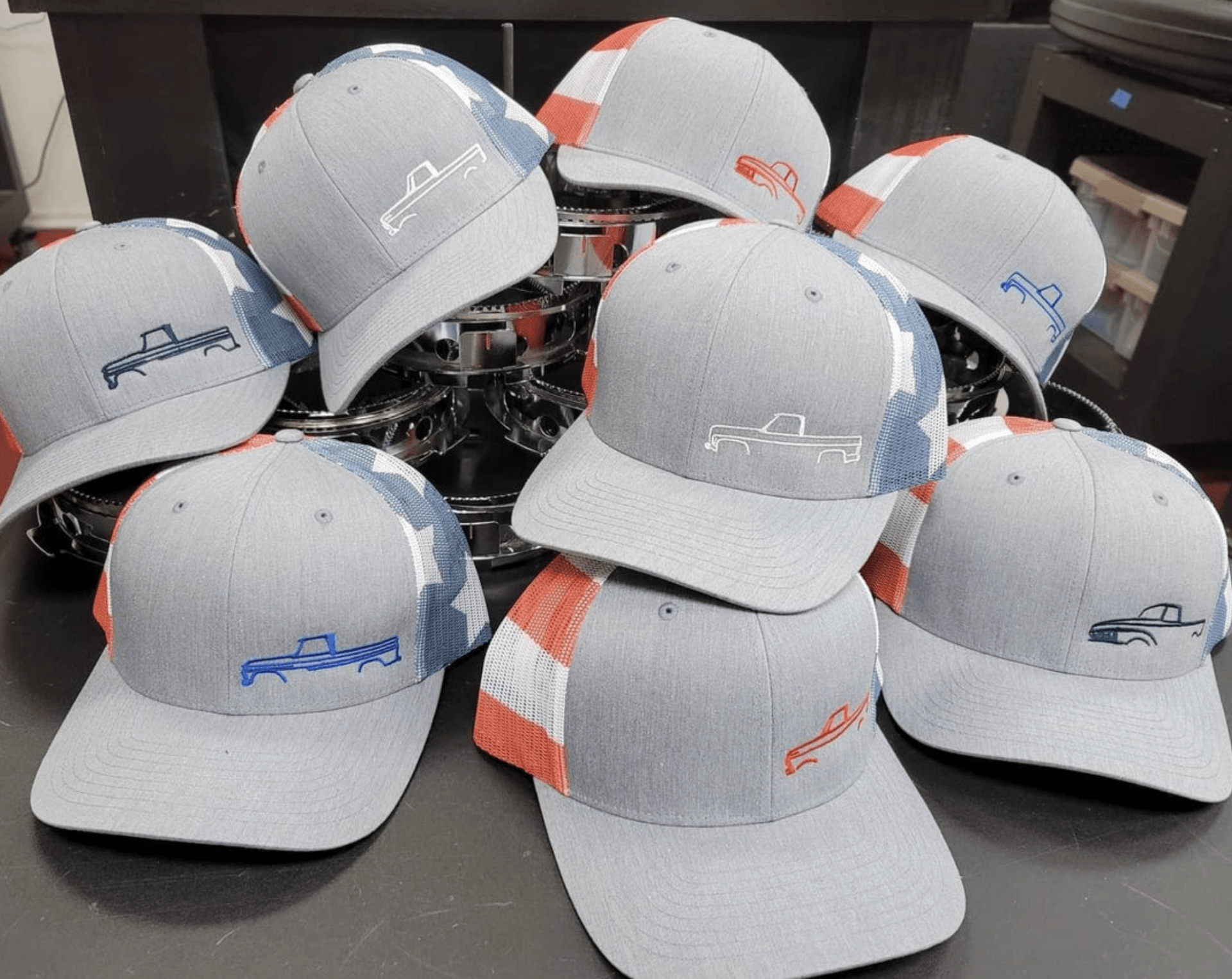 Eight gray trucker hats with red and white accents, embroidered with a logo and a word on the front.