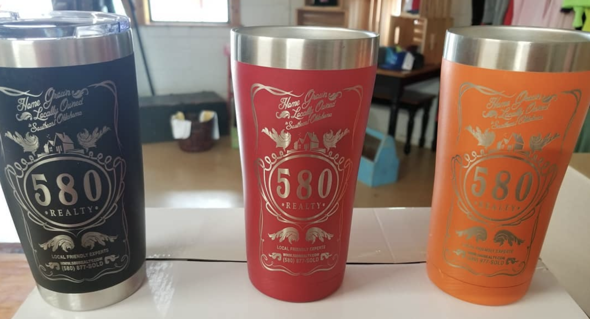 Three insulated tumblers: black, red, and orange, with silver rims and a gold logo.