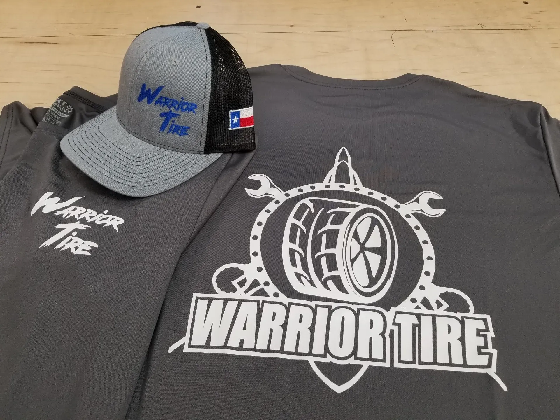 Gray Warrior Tire hat and shirts with white logos on a wood surface.