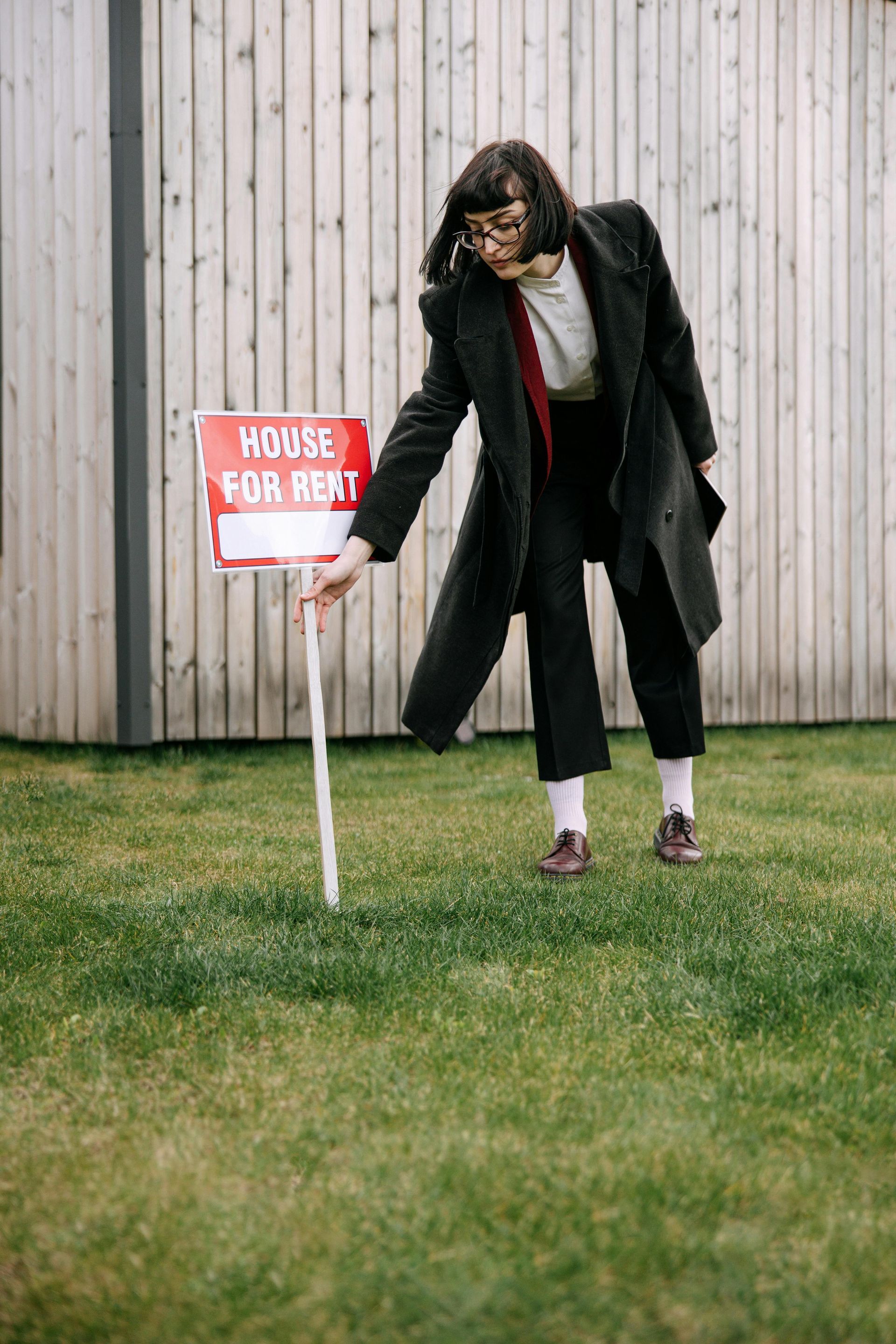 A woman is pointing at a house for rent sign in the grass.