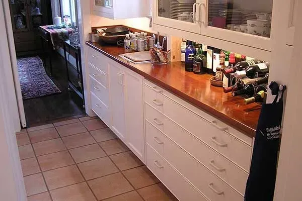 Narrow kitchen with white cabinets, wood countertop, and tiled floor, viewed down a hallway.