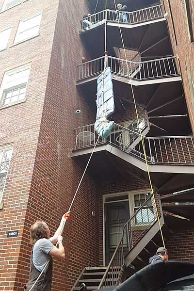 Workers lower a bundle down an exterior brick stairwell with ropes on an apartment building