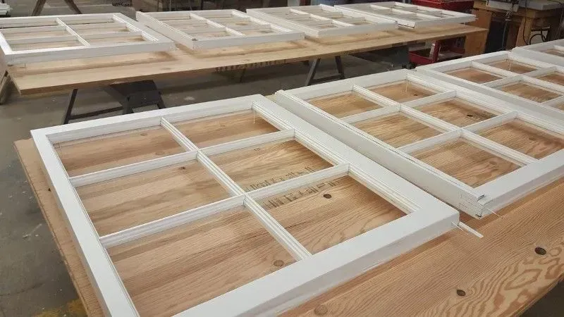 White-framed wooden window sashes lined up on a workshop bench during construction