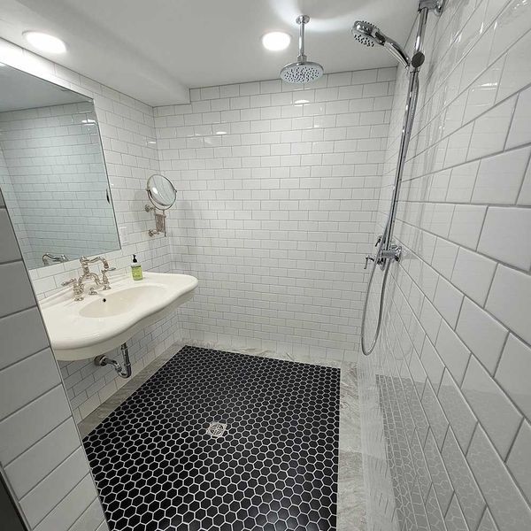 Small white-tiled bathroom with black hex tile floor, pedestal sink, mirror, and walk-in shower.