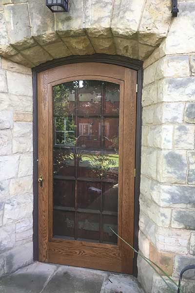 Wooden arched door with glass panes set in a stone wall, reflecting greenery outside.