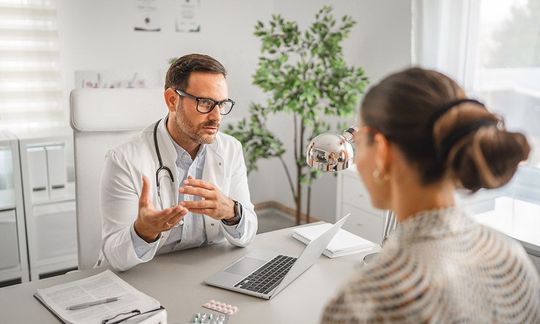 A doctor wearing a white coat and stethoscope talks to a patient across a desk in a bright, modern medical office.