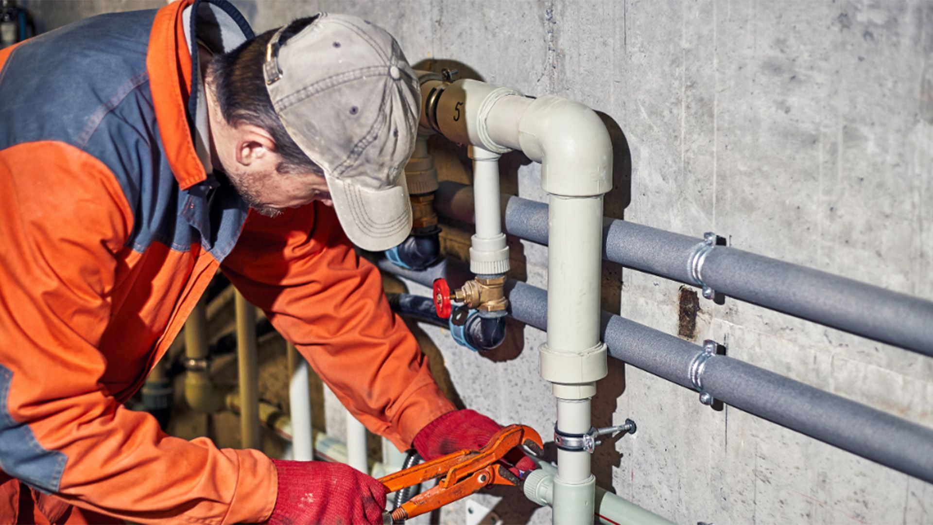 Plumber in orange jacket and red gloves working on pipes with pliers in a utility room. Plumber in orange jacket and red gloves working on pipes with pliers in a utility room.