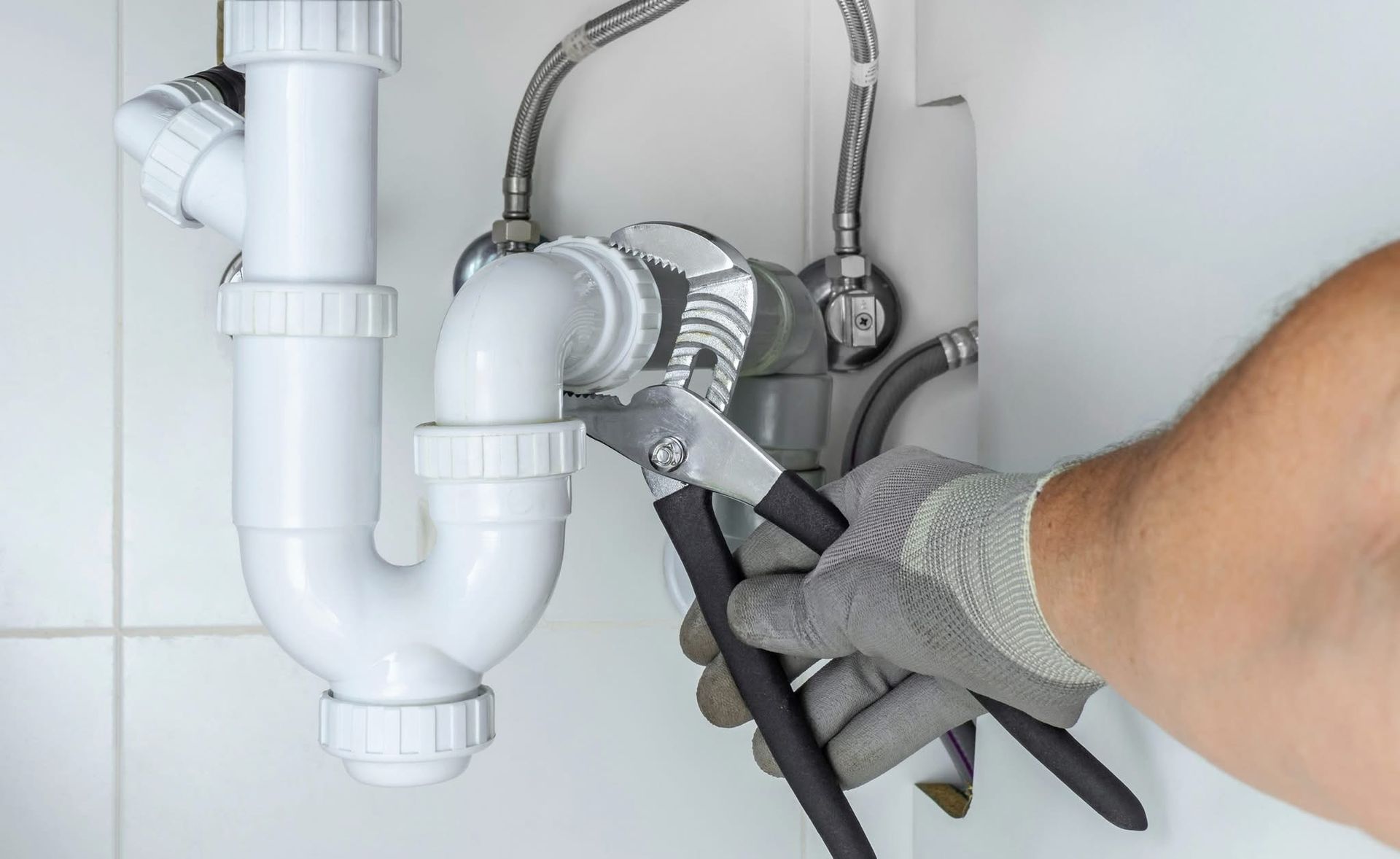 A plumber uses pliers to tighten a white plastic pipe under a sink. A plumber uses pliers to tighten a white plastic pipe under a sink.