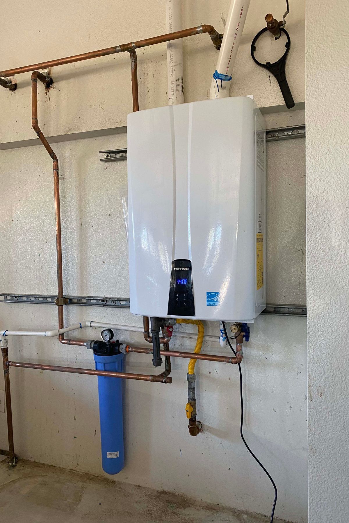 White tankless water heater with copper pipes and blue water filter on a concrete wall. White tankless water heater with copper pipes and blue water filter on a concrete wall.
