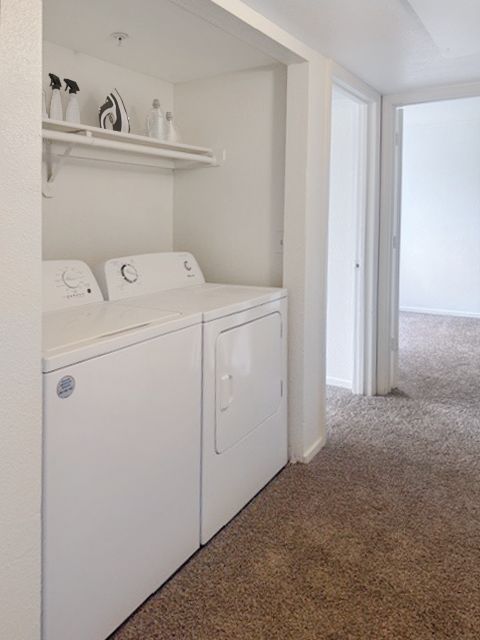 White washer and dryer in a carpeted laundry nook with an overhead shelf holding cleaning supplies.