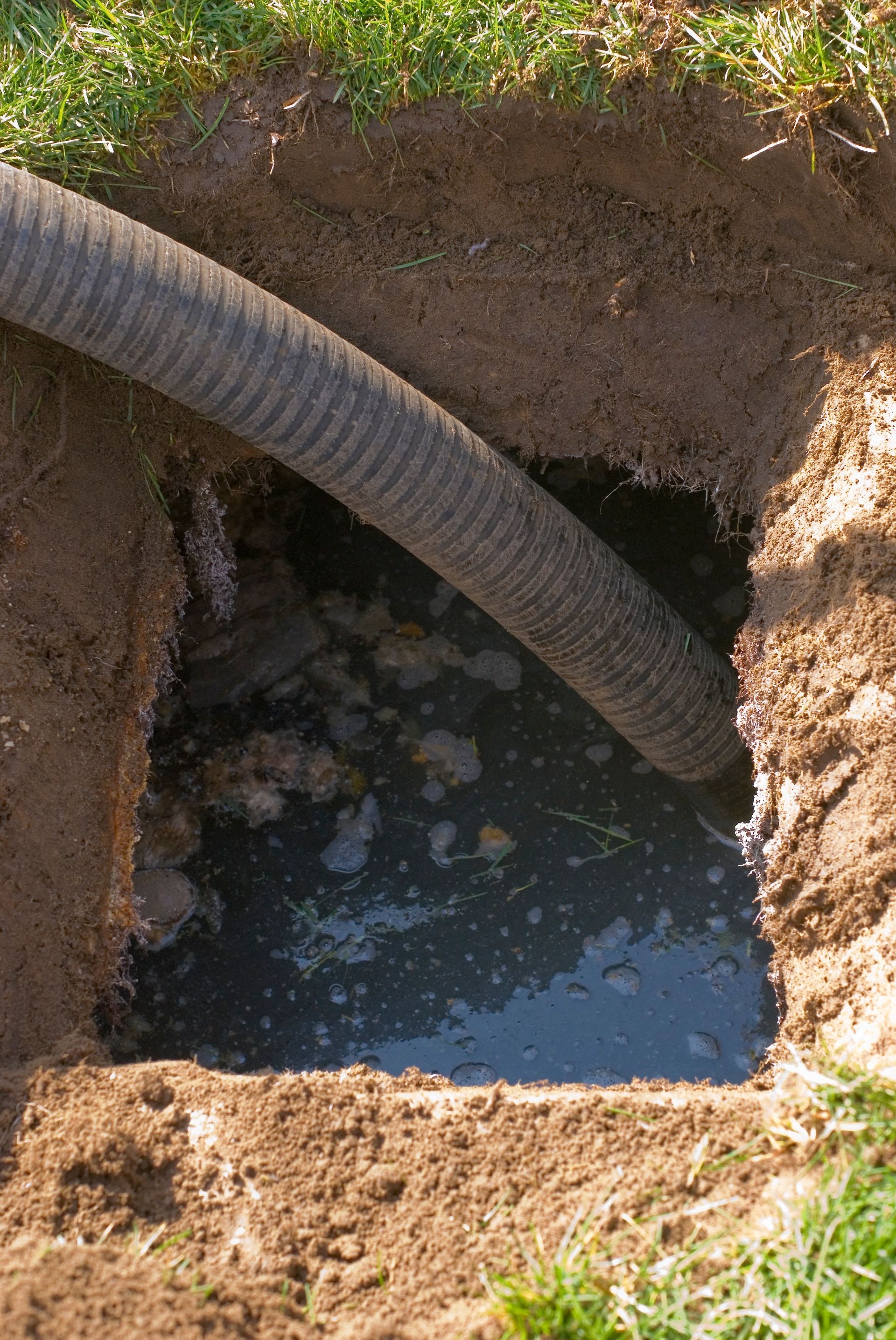 Tubo che drena acqua torbida da una buca quadrata scavata nel terreno, circondata dall'erba.