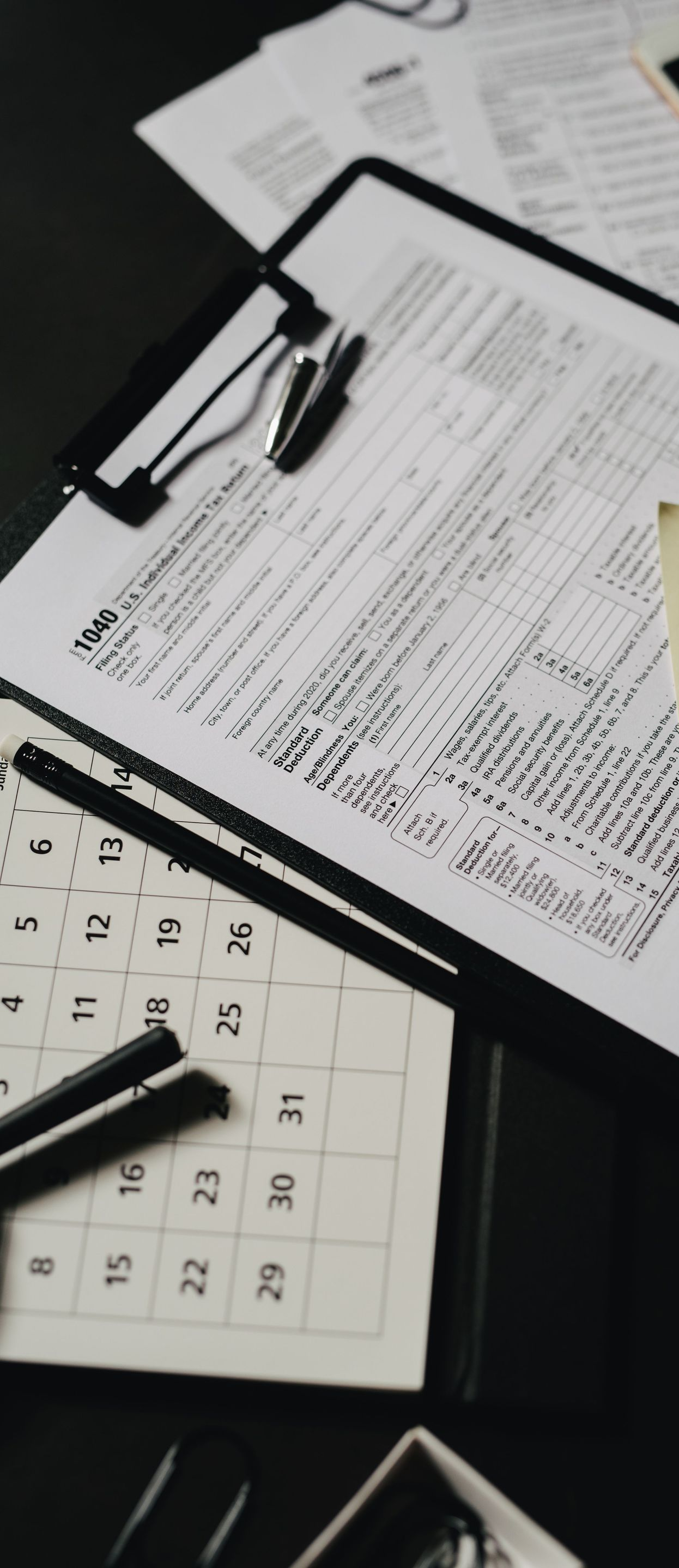 Hands writing in a notebook beside a red plaid notebook and a wooden desk with bills and papers