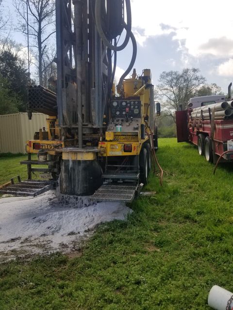 Drilling rig operating on a grassy site, with a worker in a yellow hard hat beside it.