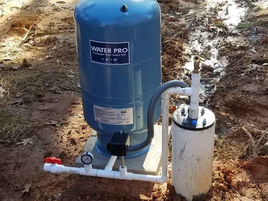 Blue well pump and white pressure tank connected by pipes in a dirt yard