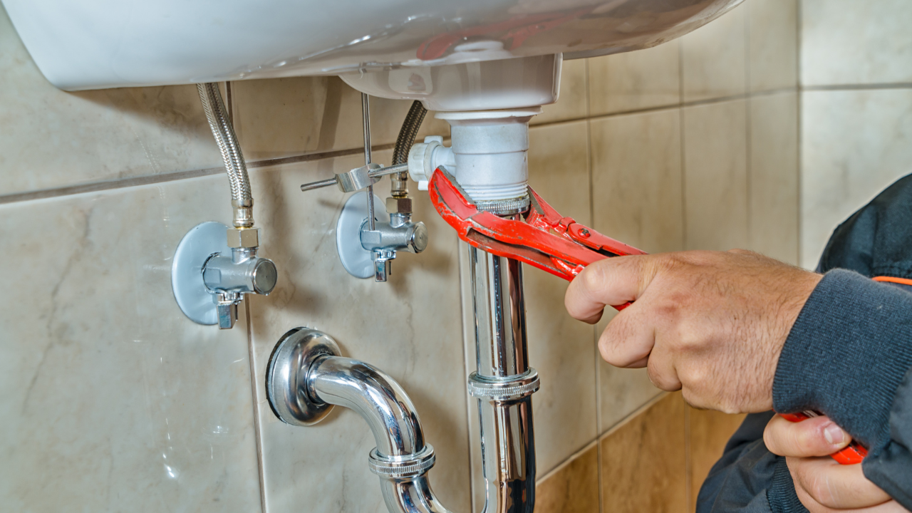 Plumber using a wrench to work on pipes under a white sink in a tiled bathroom.