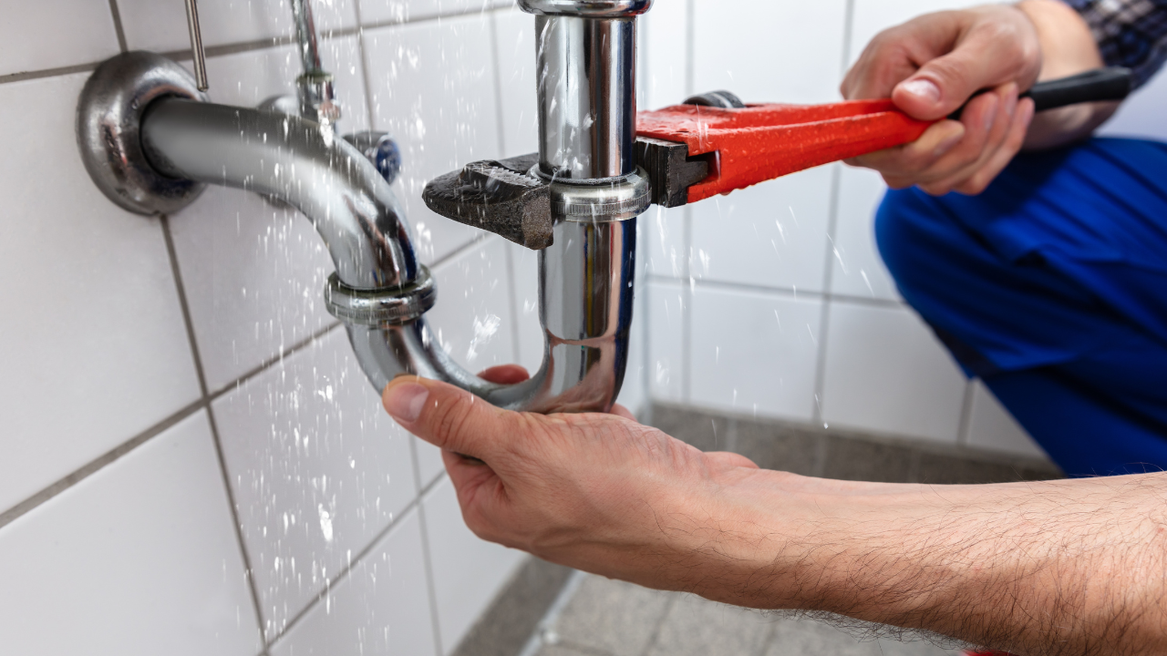 Plumber using a wrench on a leaking chrome sink pipe in a tiled bathroom.