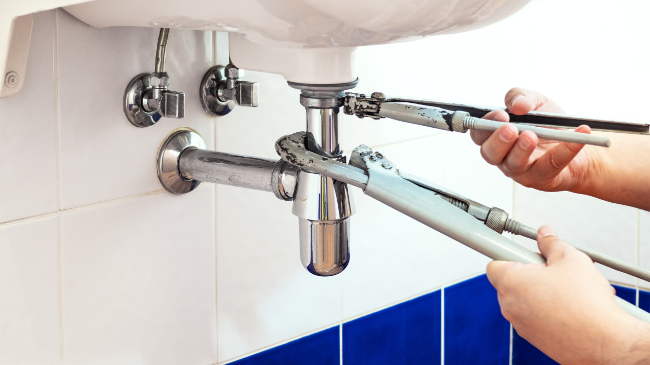 Hands using wrenches to tighten or loosen plumbing under a white sink.