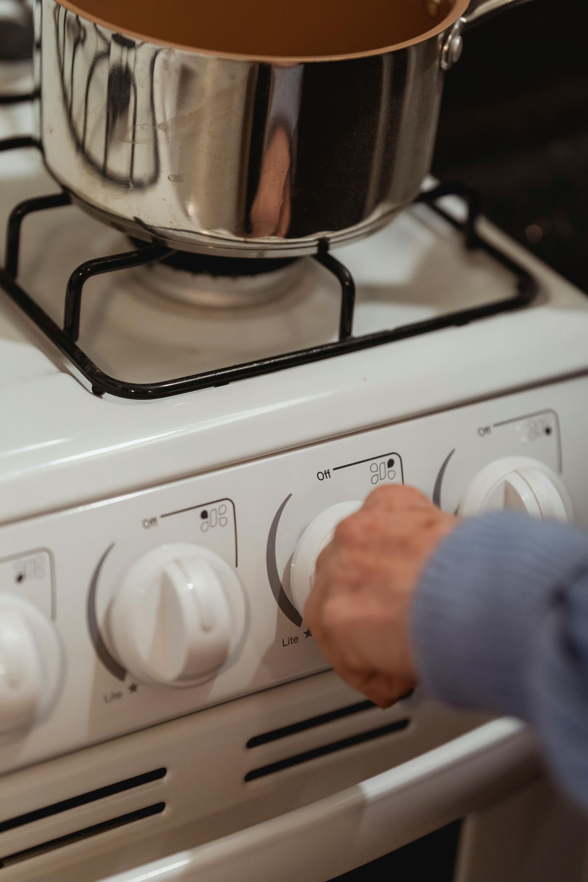 A hand turns a stove knob, with a pot on the burner.