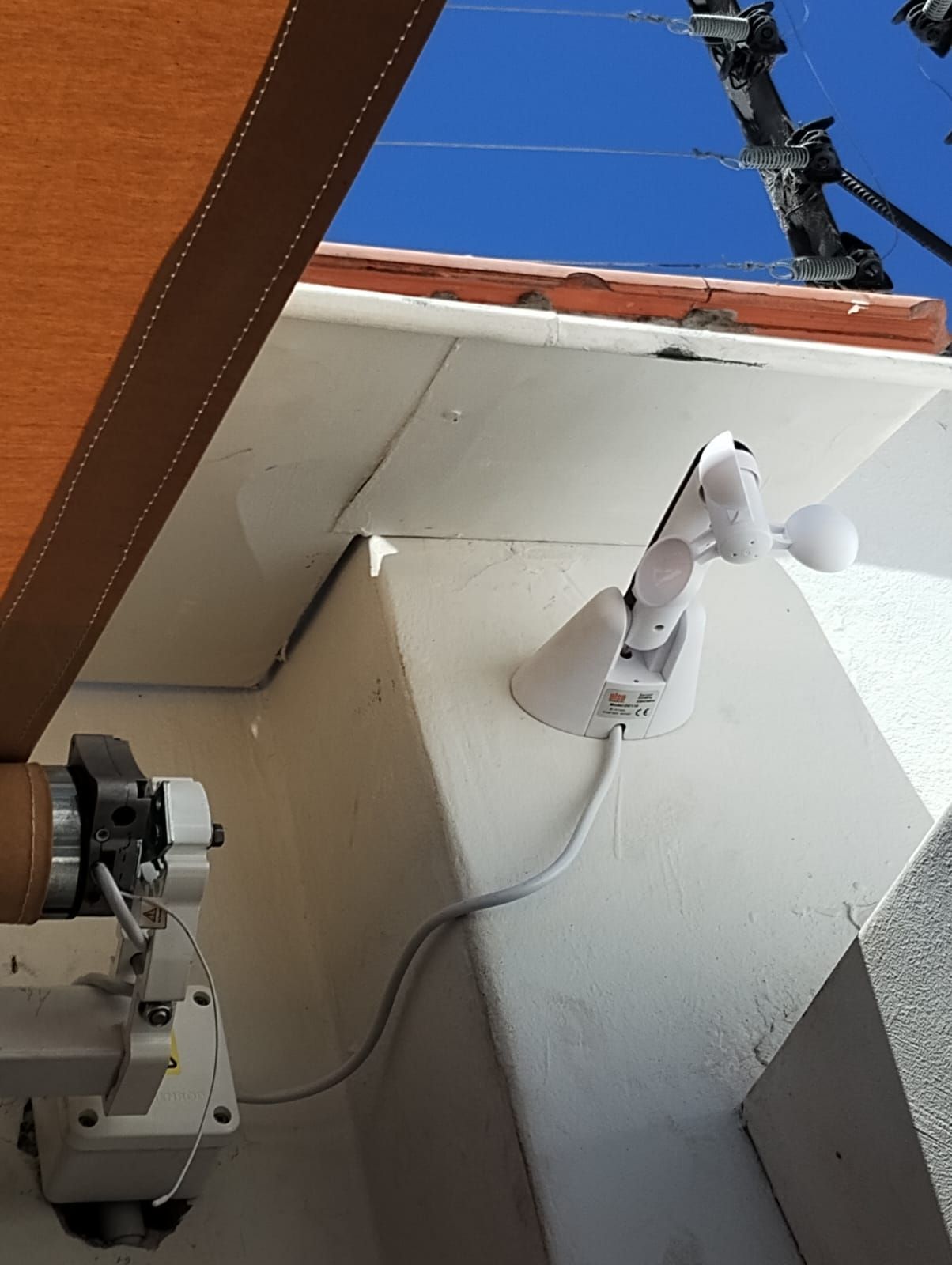 Anemómetro blanco en una esquina de un edificio, cable bajando, toldo marrón, cielo azul.