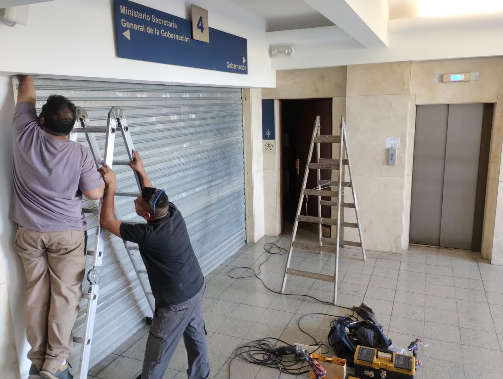 Dos hombres instalan una persiana metálica de seguridad en un edificio. Se utiliza una escalera.