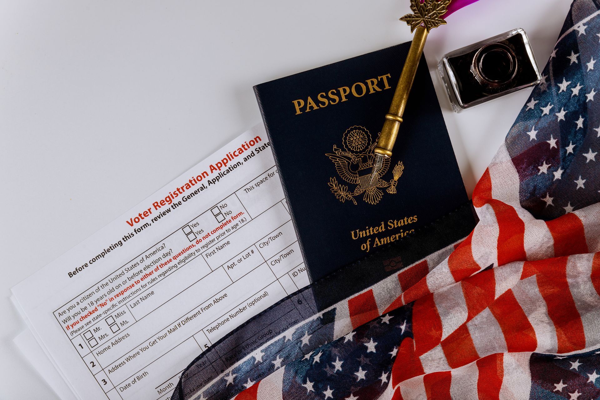 US passport, application form, quill pen, inkwell, and American flag on a white surface.