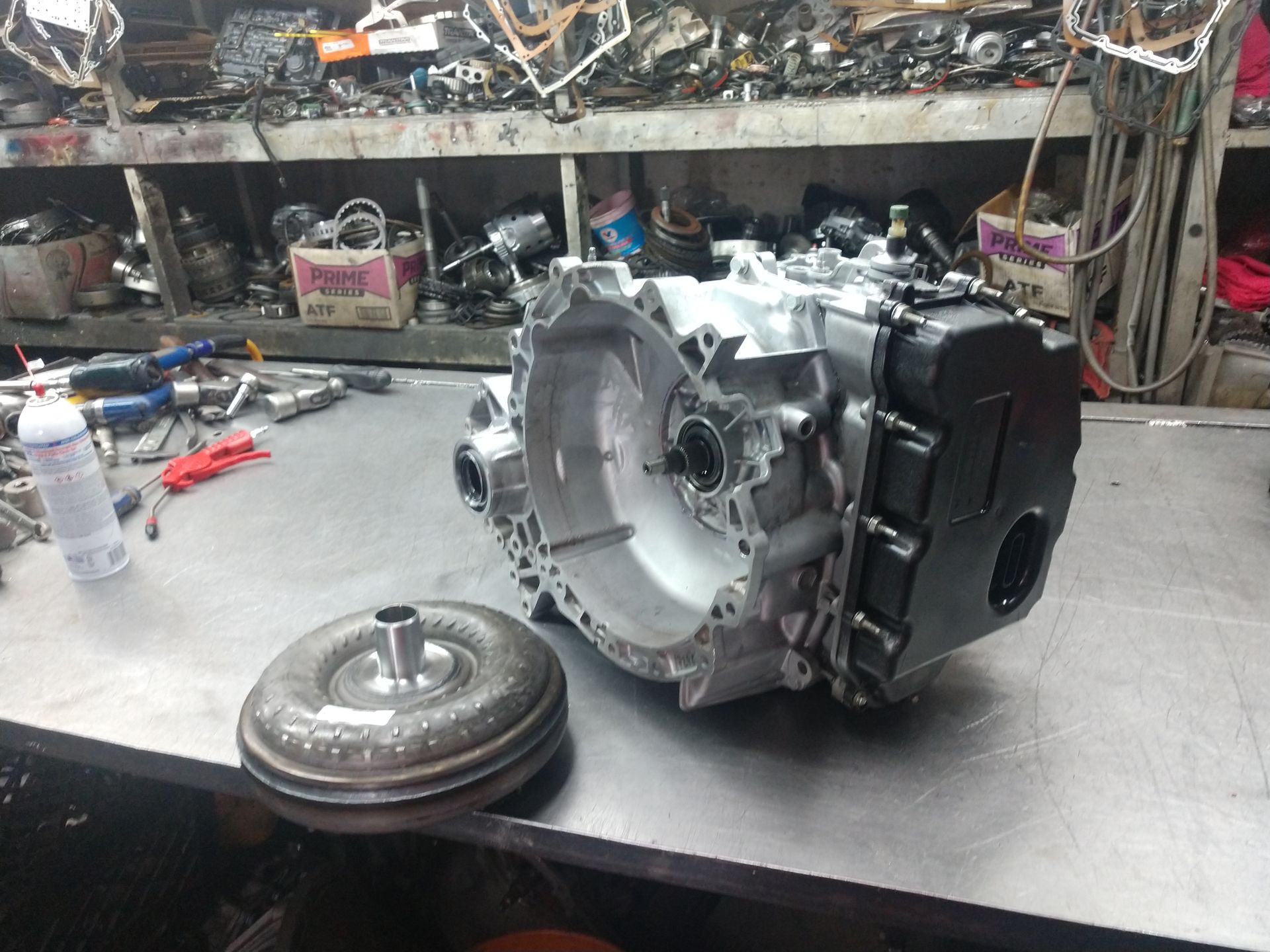 Car transmission housing on a workbench in a garage, with a torque converter and tools nearby