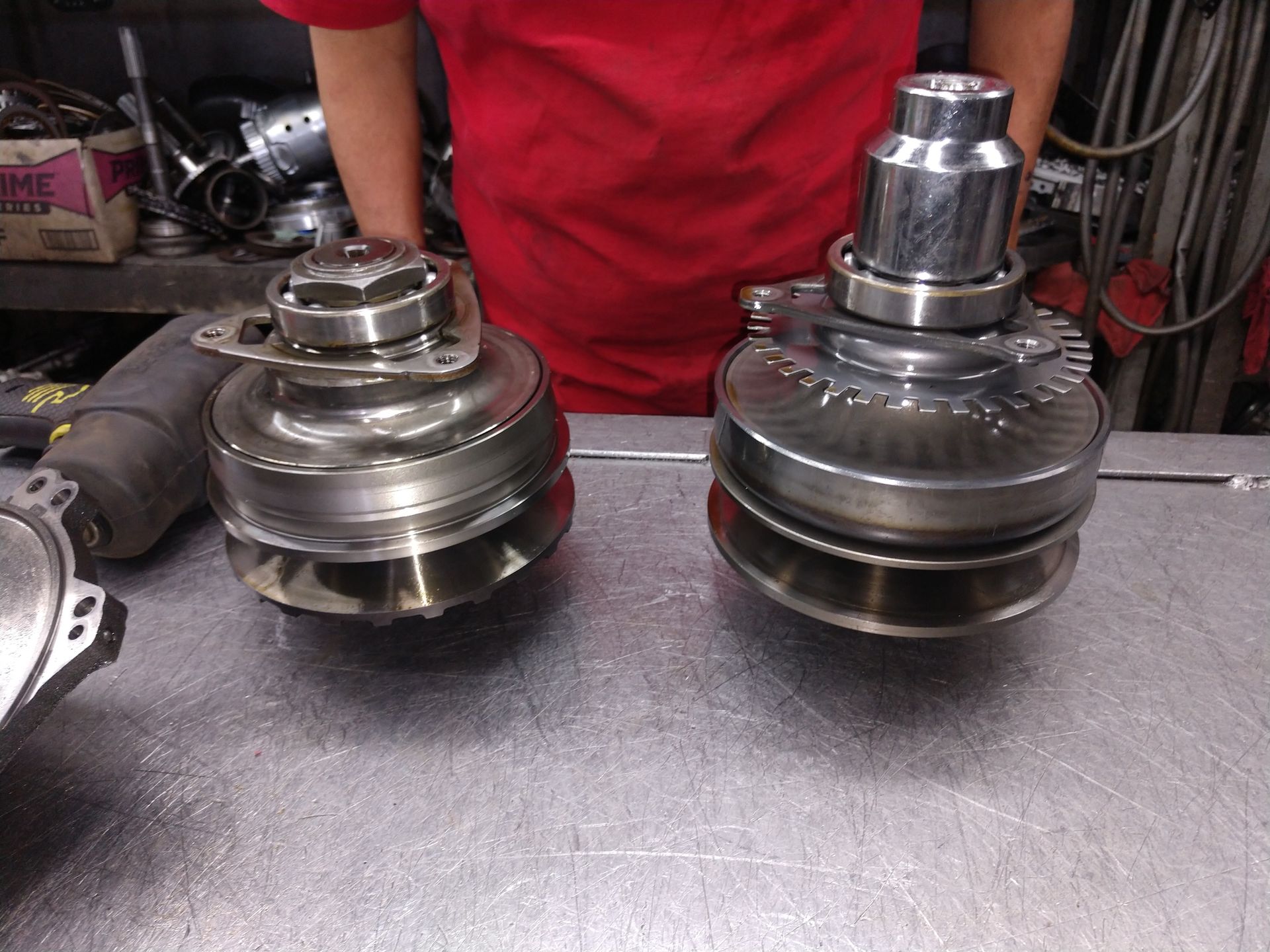 Two metal automotive parts on a workbench, with a person in a red shirt standing behind them.