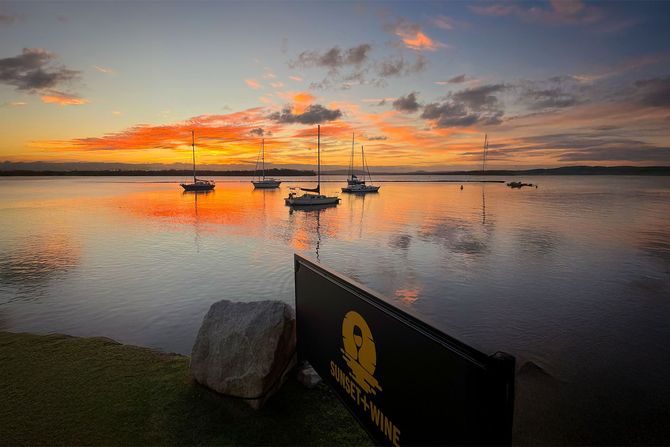 Sunset over calm water with sailboats; orange and yellow sky reflected in the water.