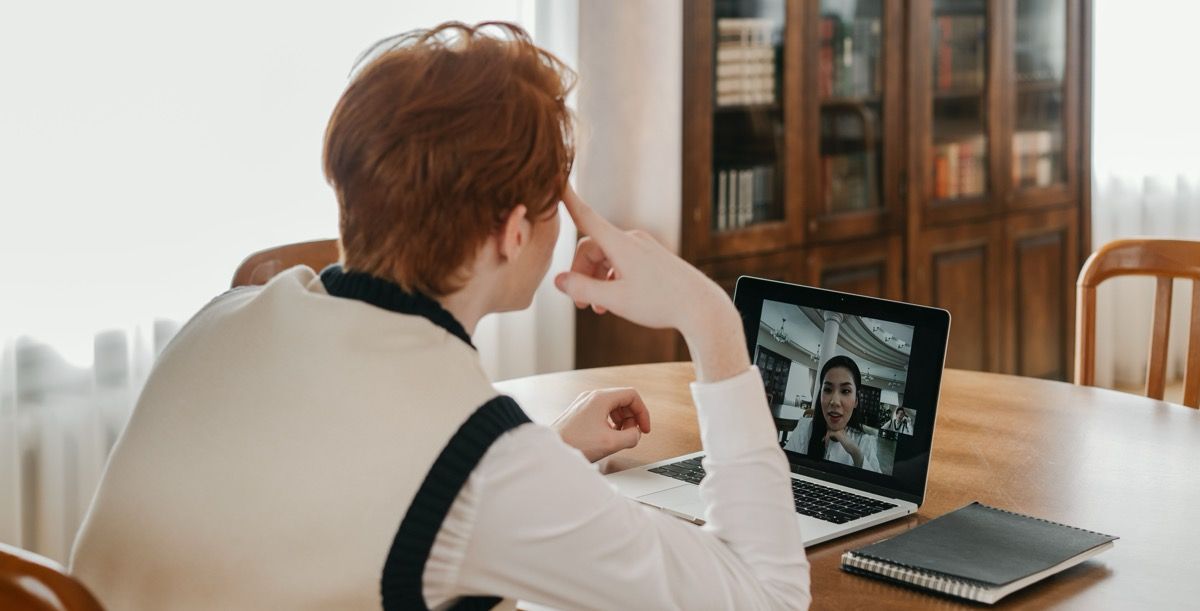 Person on a video call at a dining table, using a laptop in a bright room.