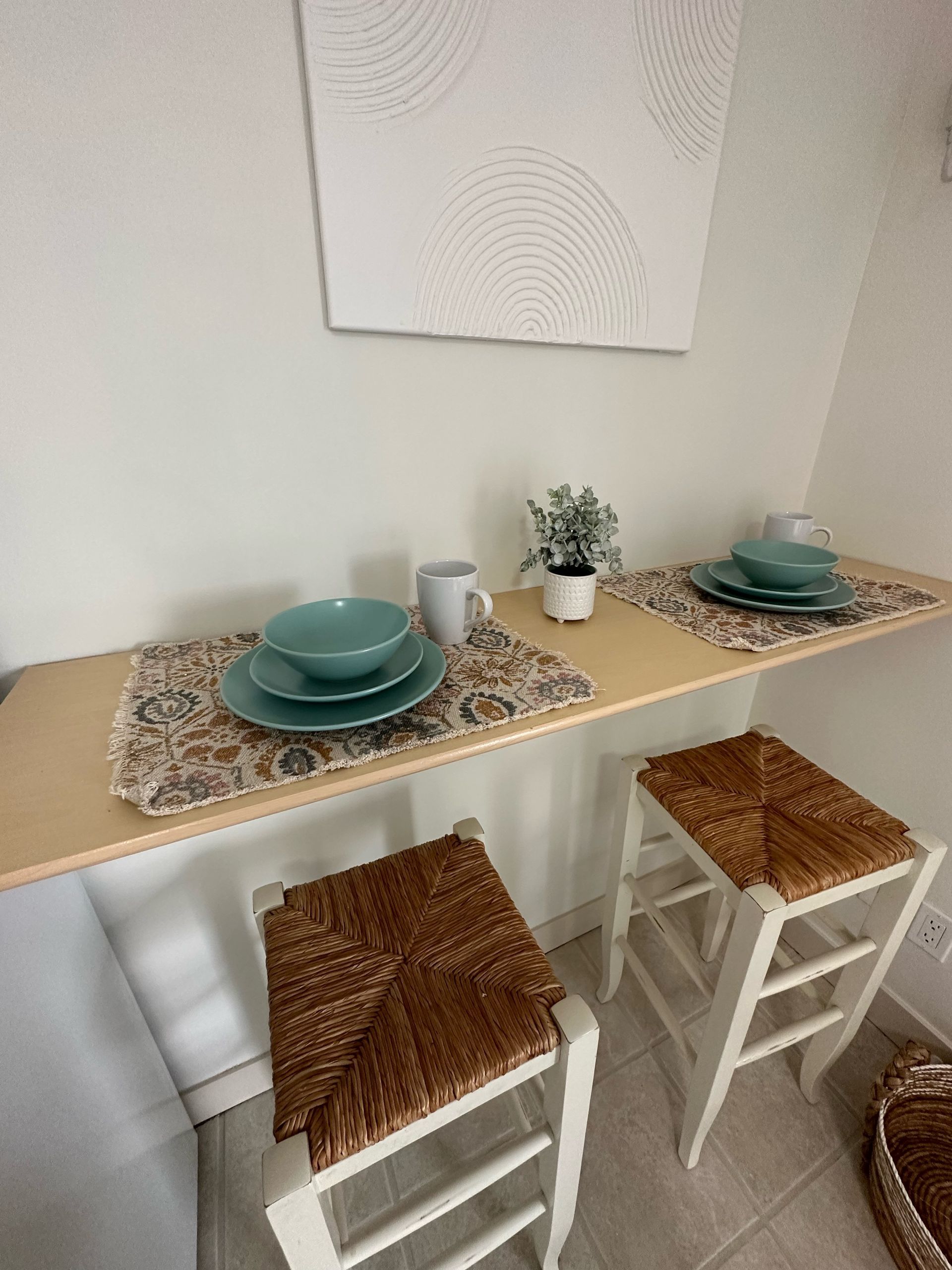 A wooden wall-mounted bar table with two wicker-topped stools, place settings, and a small plant against a white wall.