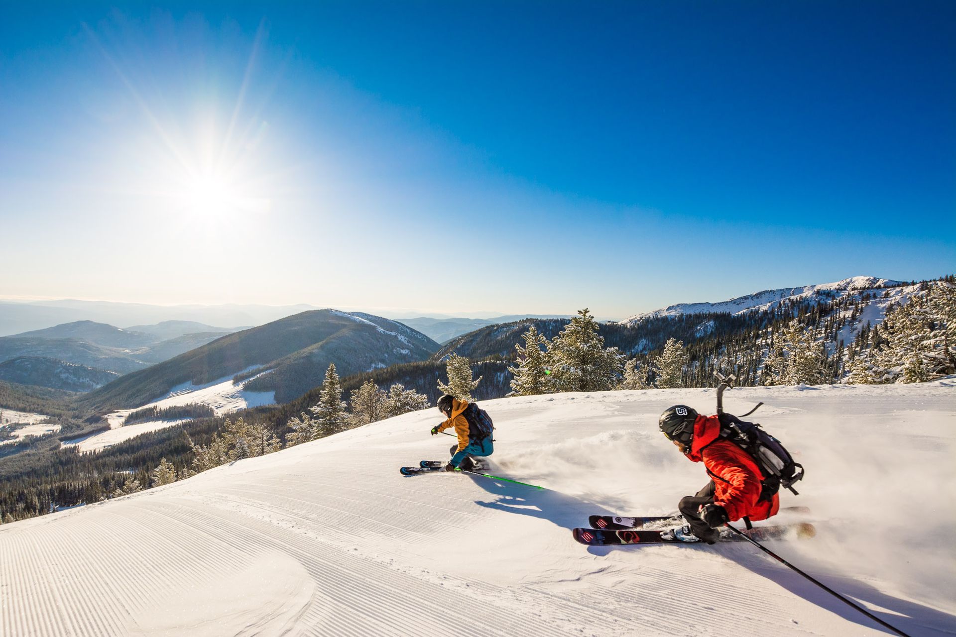 Two skiers carve down a sunny, snow-covered mountain slope, with distant snow-capped peaks under a clear blue sky.