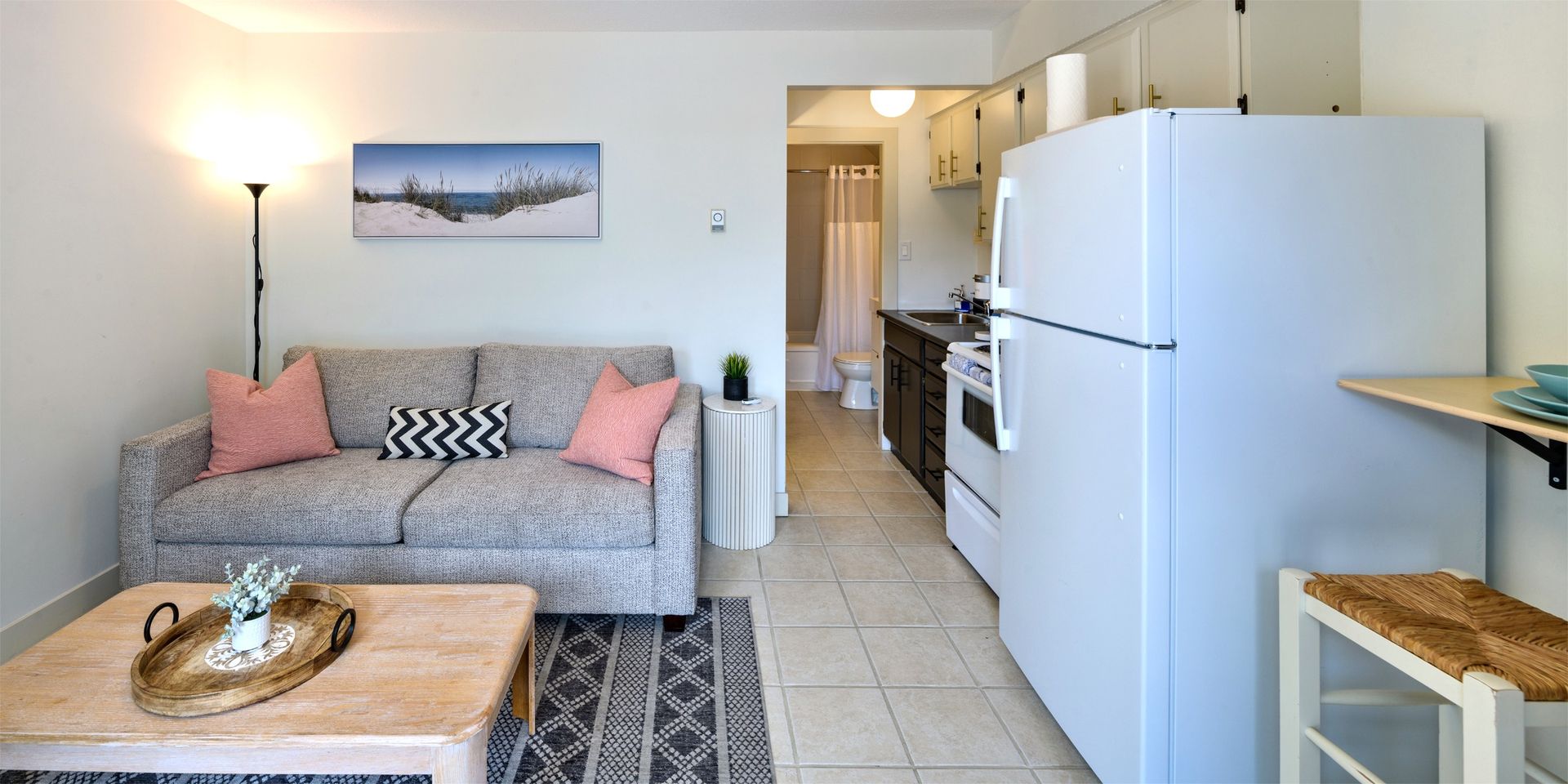 A studio apartment with a light gray sofa, two coral pillows, a wooden coffee table, a white refrigerator, and a kitchen.