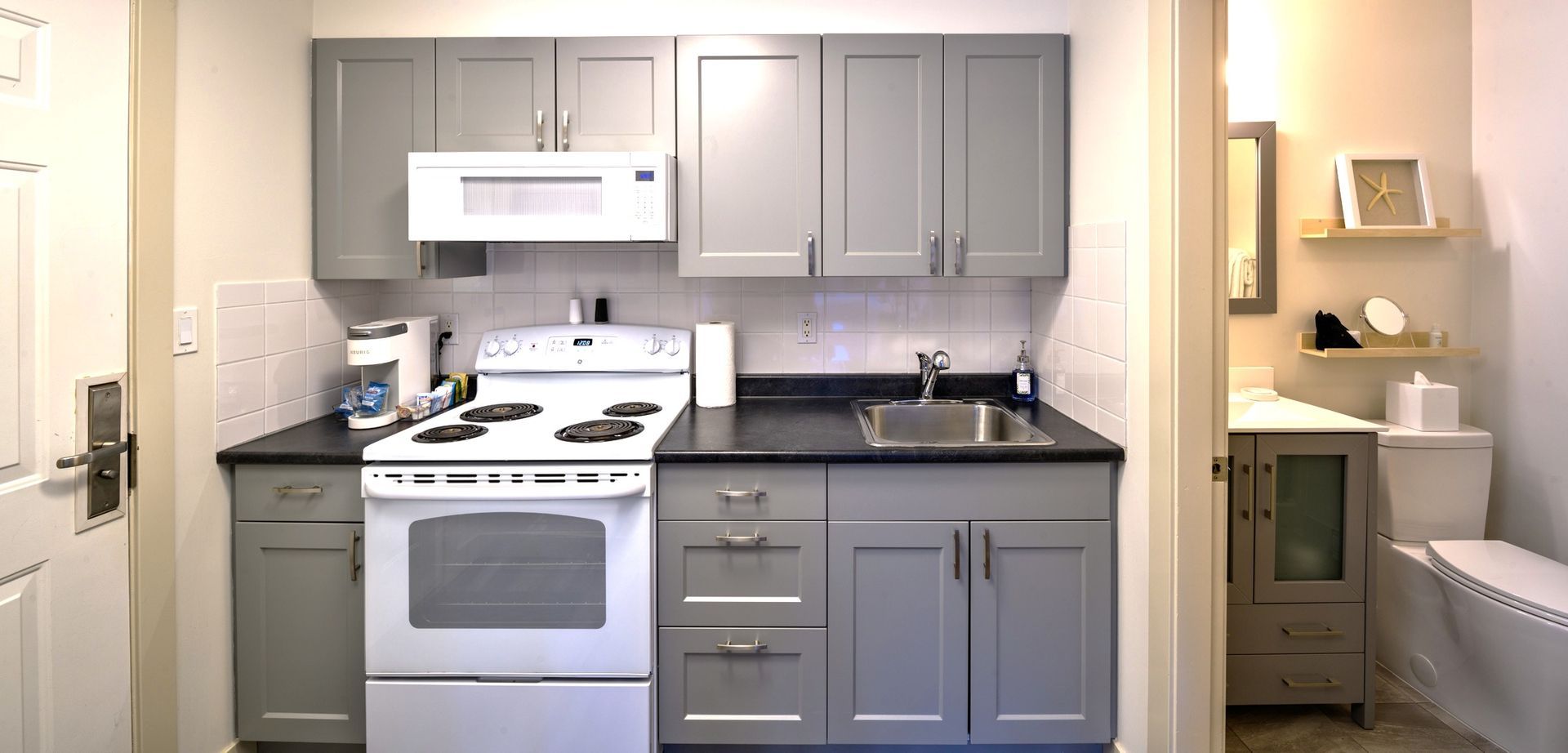 A small kitchen with gray cabinets, black countertops, and a white stove, leading into a bathroom with a white toilet.