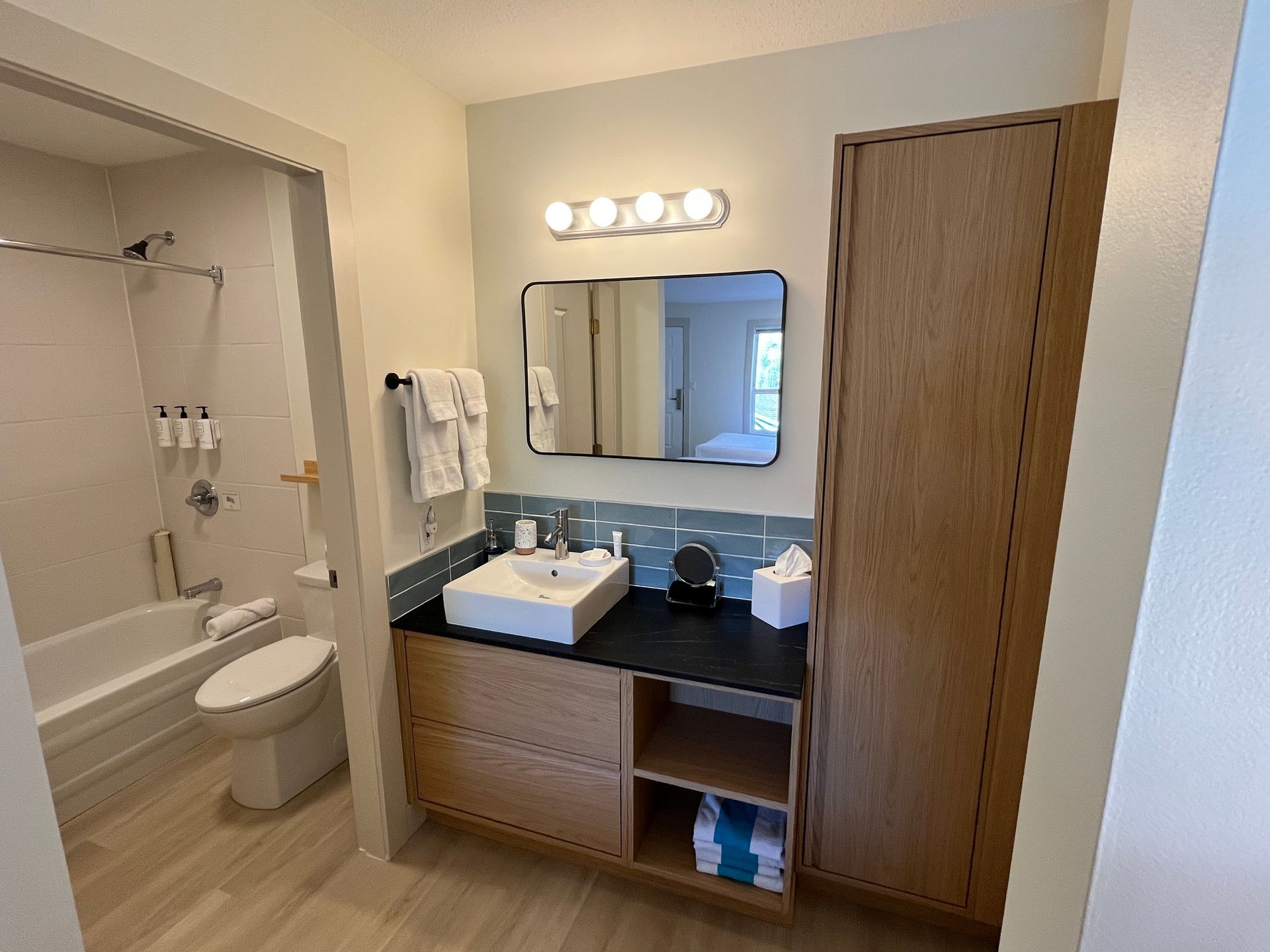 A modern hotel bathroom featuring a vanity with a wood cabinet, black countertop, rectangular mirror, and adjacent shower.
