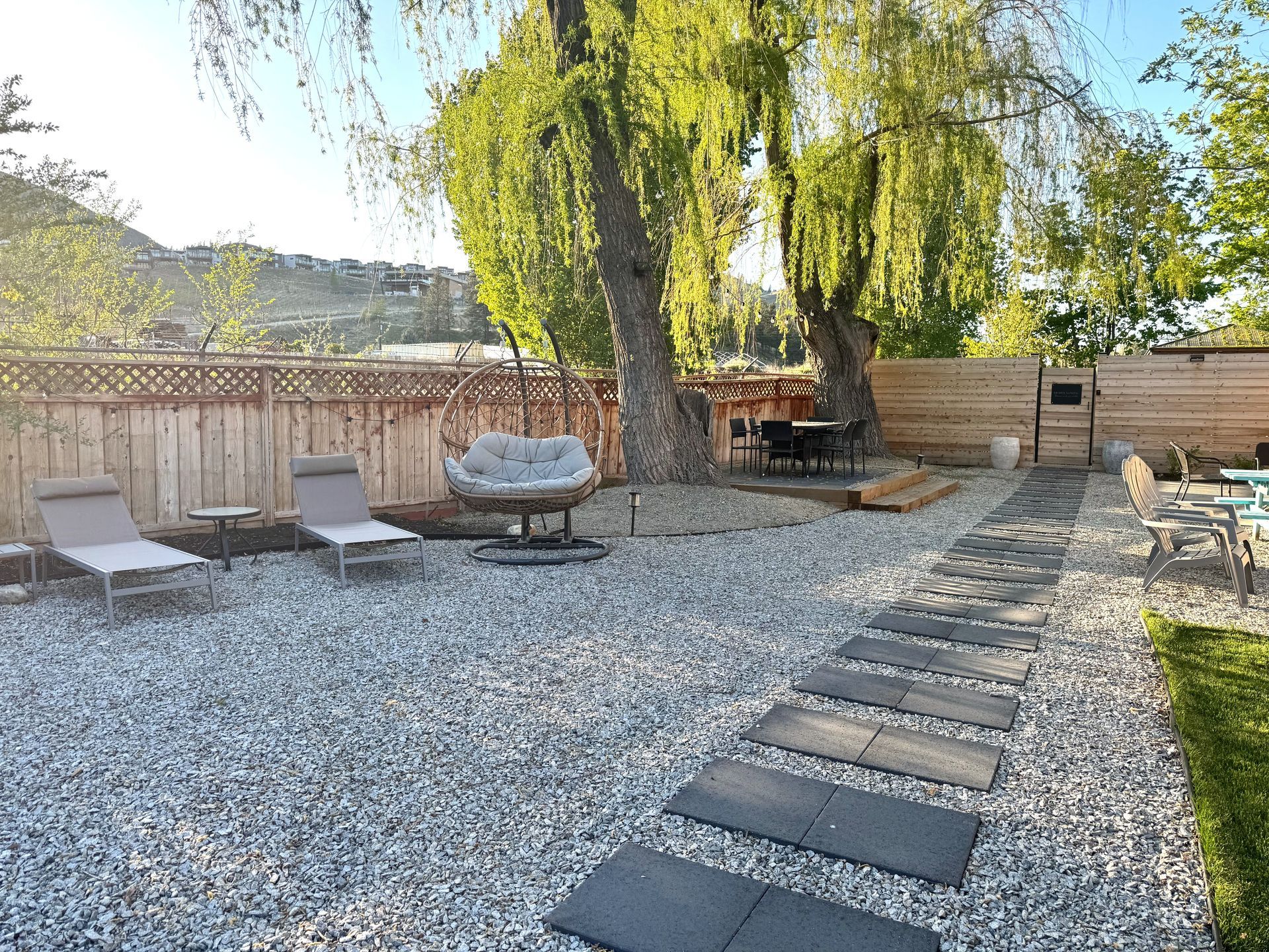 A sunny backyard with gravel ground, lounge chairs, a hanging wicker chair, a stone path, and large trees.