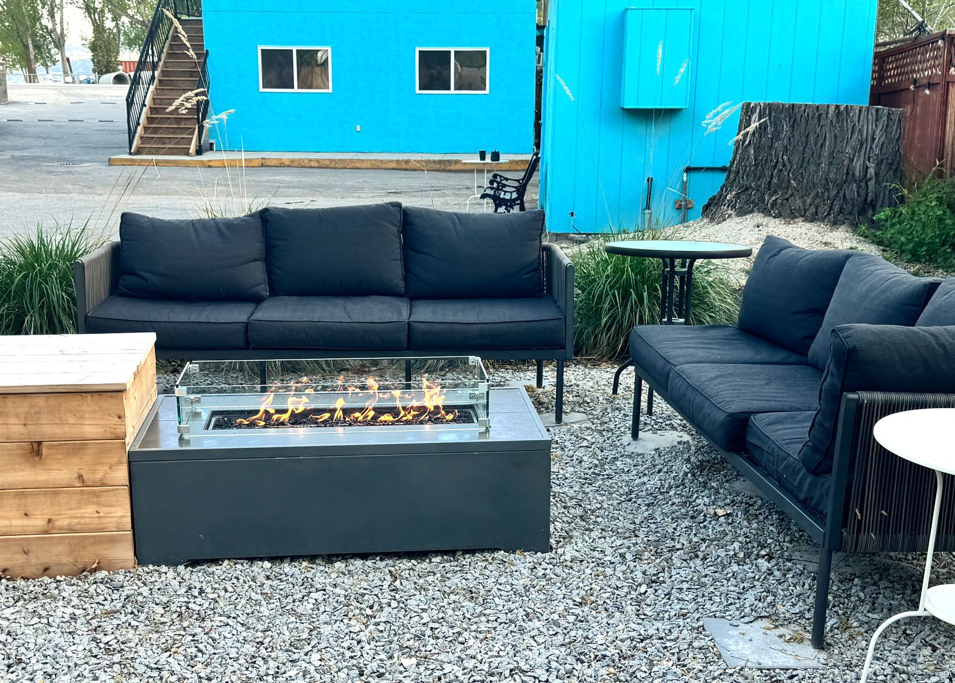 An outdoor lounge area with two dark gray sofas facing a rectangular fire pit on a gravel patio against a bright blue wall.