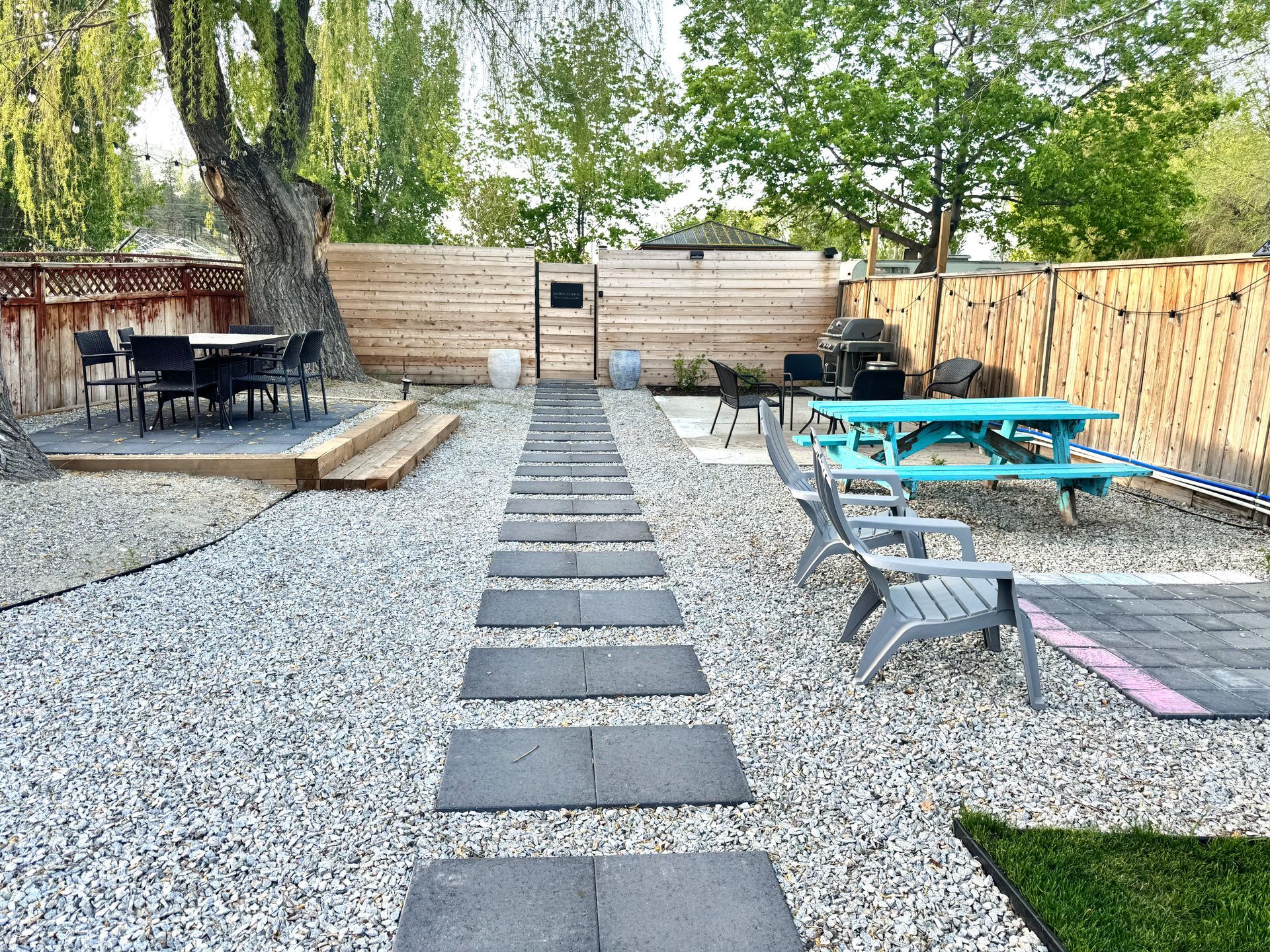 A backyard patio with a stone paver path, gravel ground, wooden fencing, a dining area, and a turquoise picnic table.