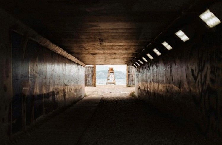 A tunnel with graffiti-covered walls looking out toward a beach with a lifeguard chair under a bright, cloudy sky.