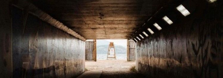 A dark tunnel with graffiti-covered walls frames a distant view of a beach and a lifeguard chair.