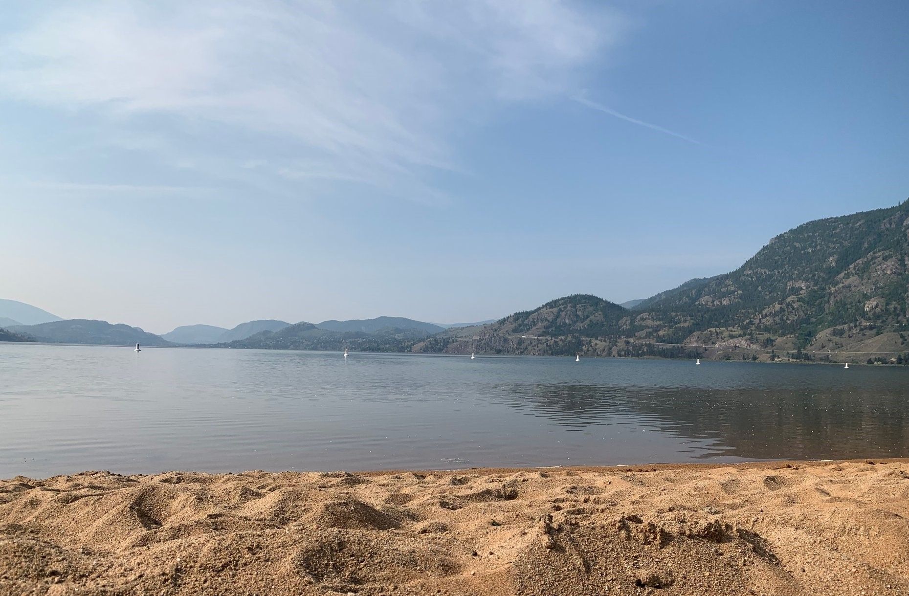 A sandy beach overlooking a wide, calm lake surrounded by hazy, forested mountains under a clear blue sky.