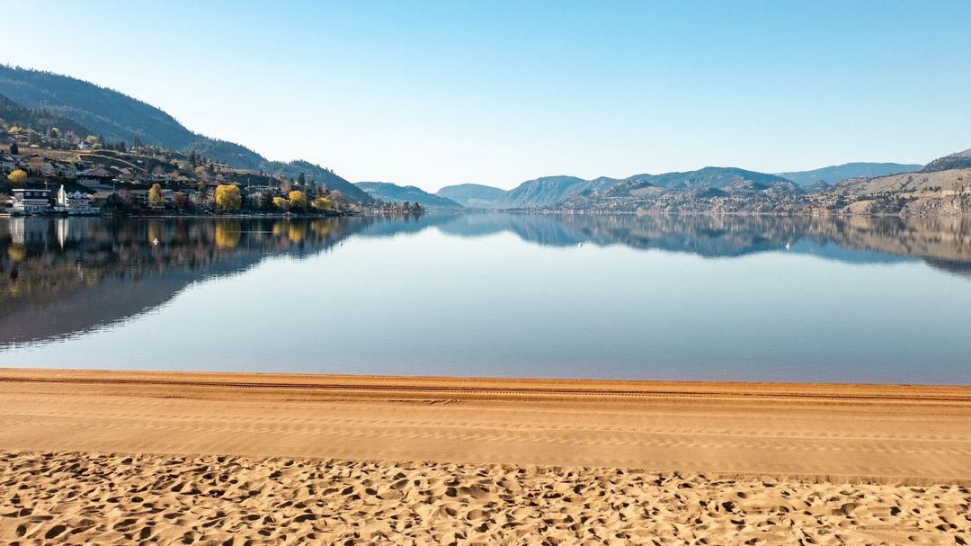 A serene, snow-covered mountain range reflects across the calm, glassy surface of a lake under a clear, pale blue sky.