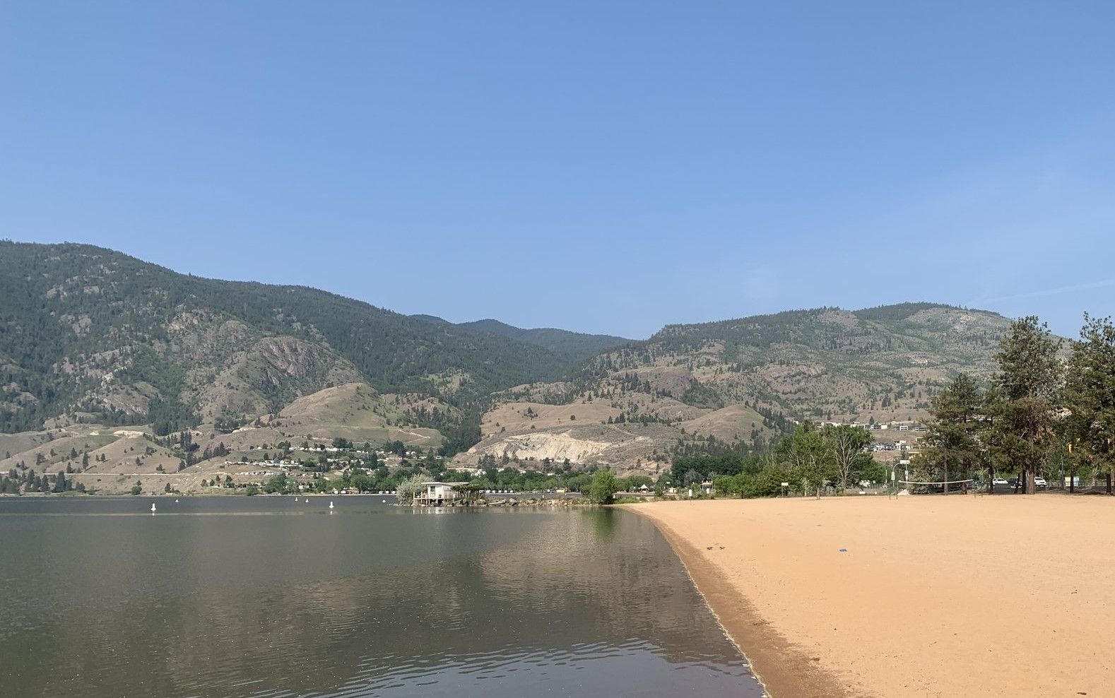 A sandy beach meets calm lake water in front of a sprawling mountain range under a clear blue sky.