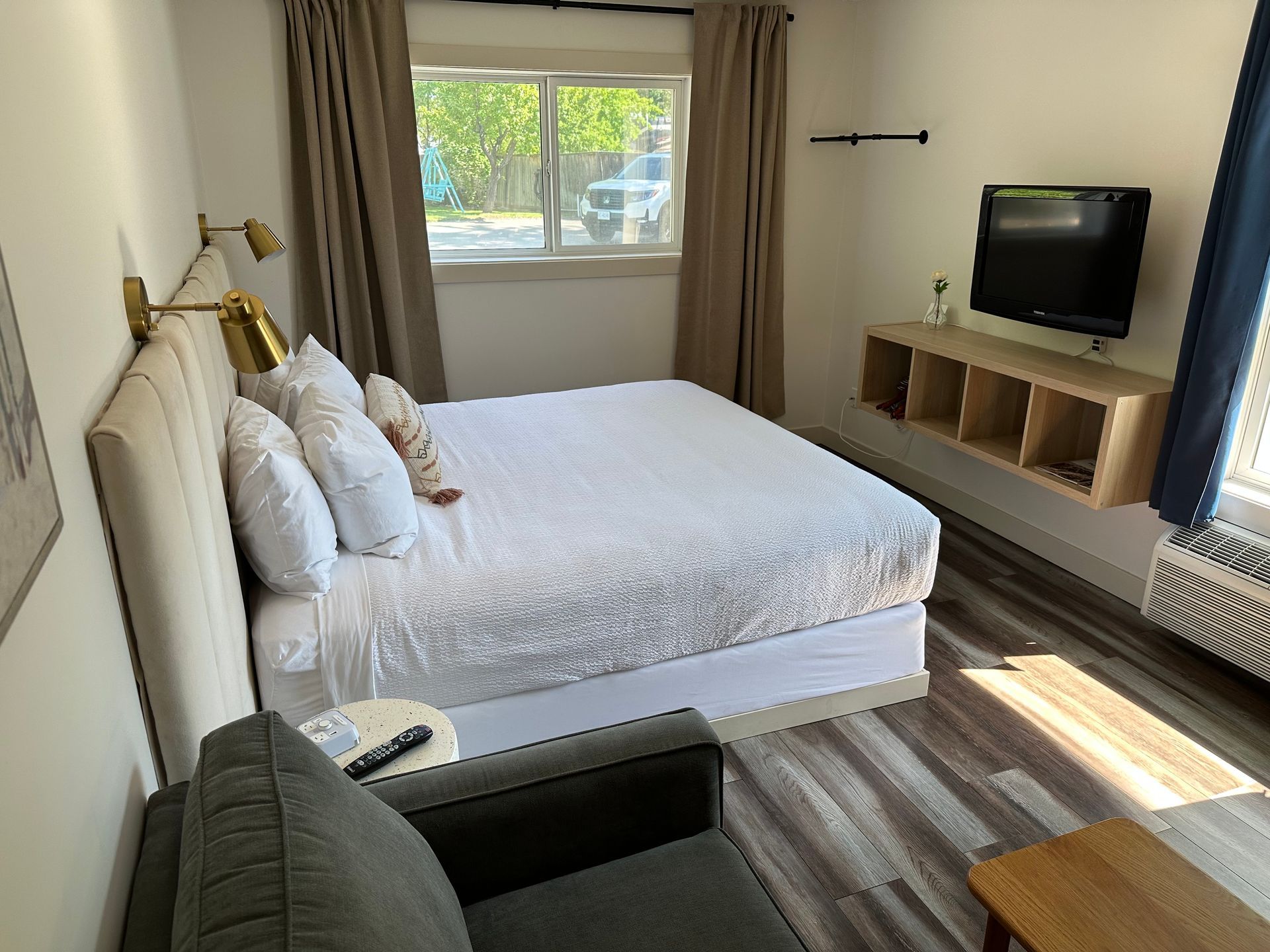 A hotel room featuring a bed with white bedding, gray chair, wall-mounted television, wood shelving, and wood-look floors.