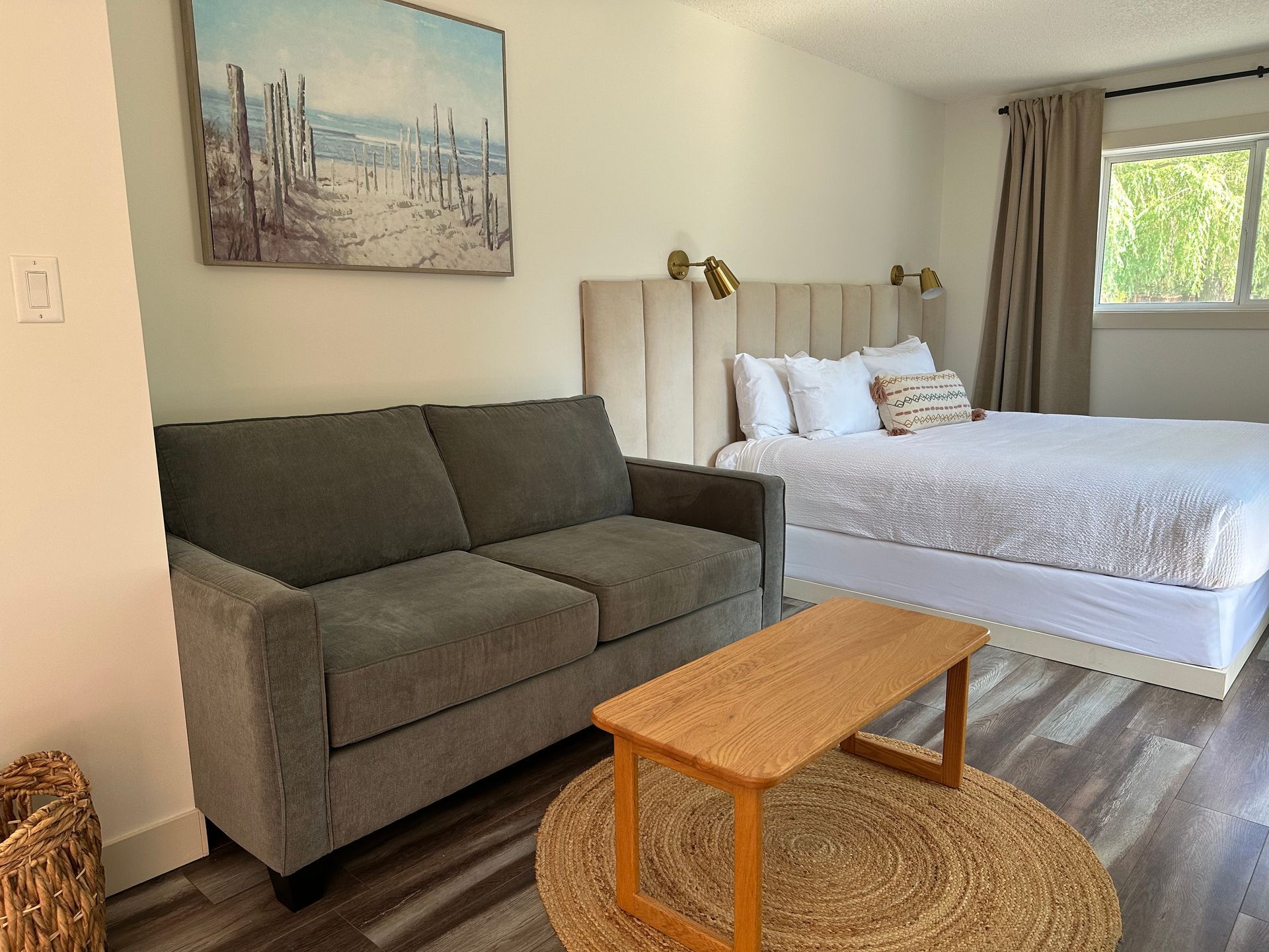 A hotel room with a grey sofa, wooden coffee table on a round rug, a queen bed with white bedding, and wall art.