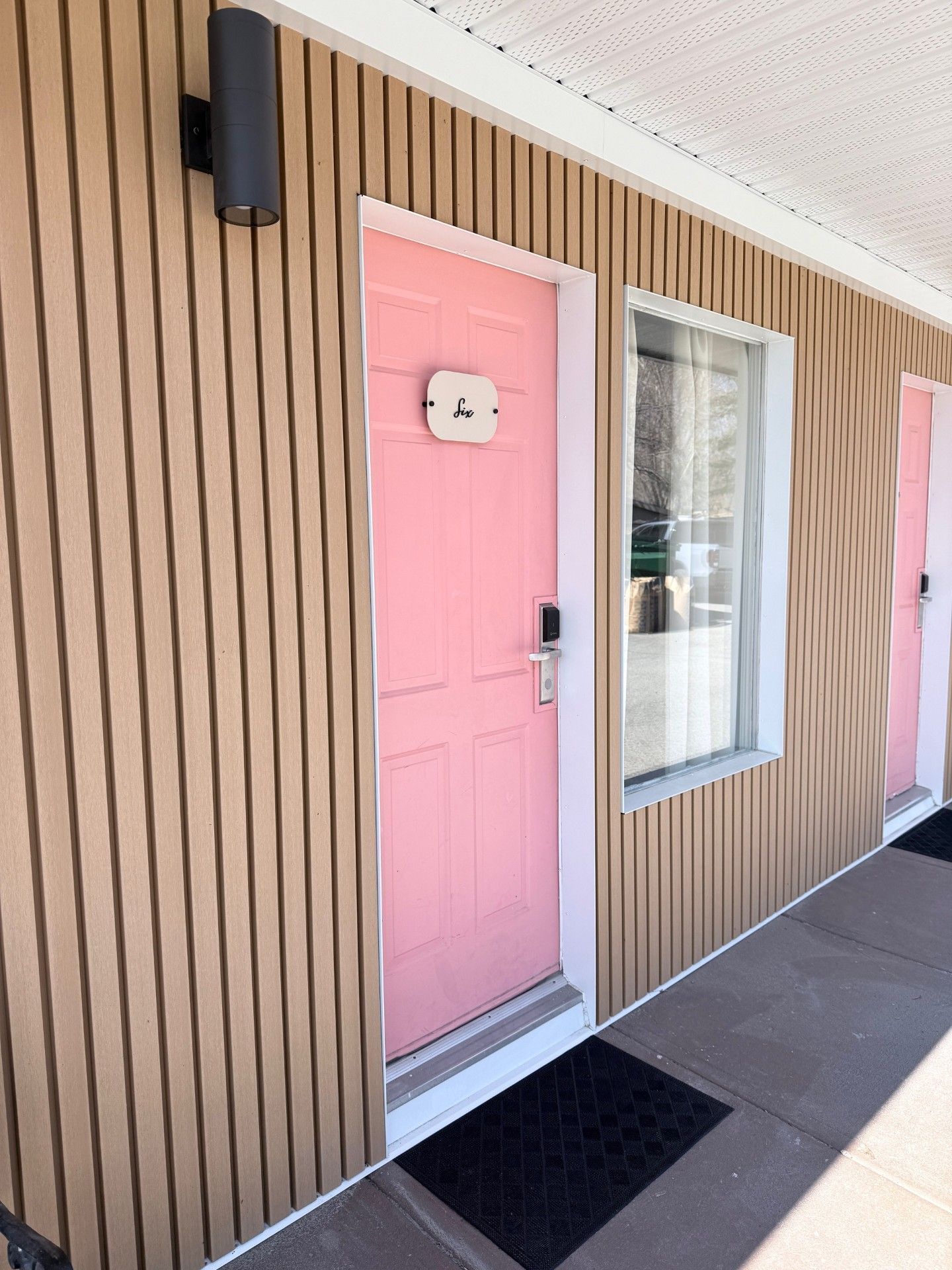 A bright pink door with a digital lock and a wall-mounted light on a building exterior with tan vertical wood siding.