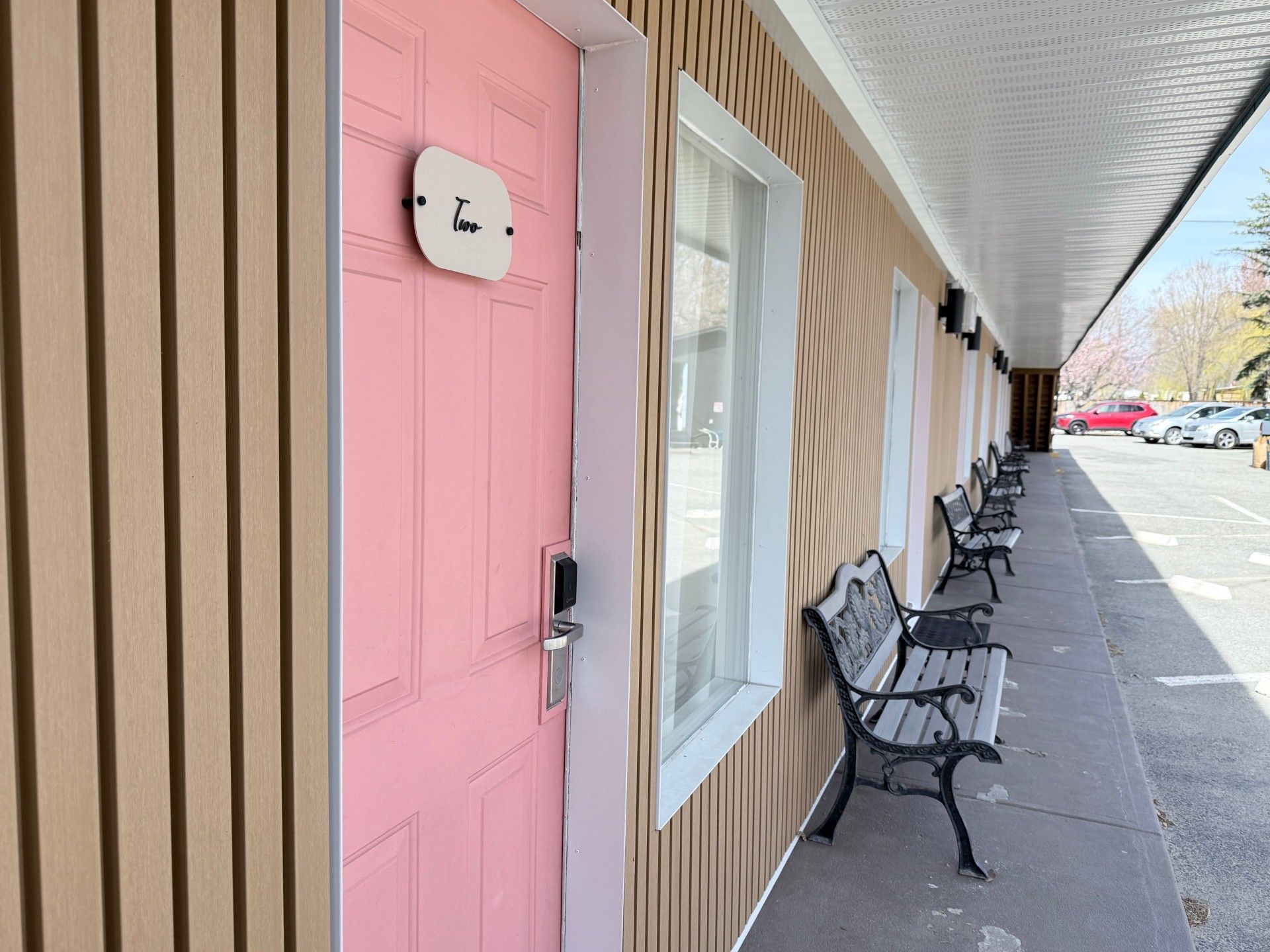 A row of motel rooms with pink doors and metal benches sits along a concrete walkway under a covered outdoor corridor.